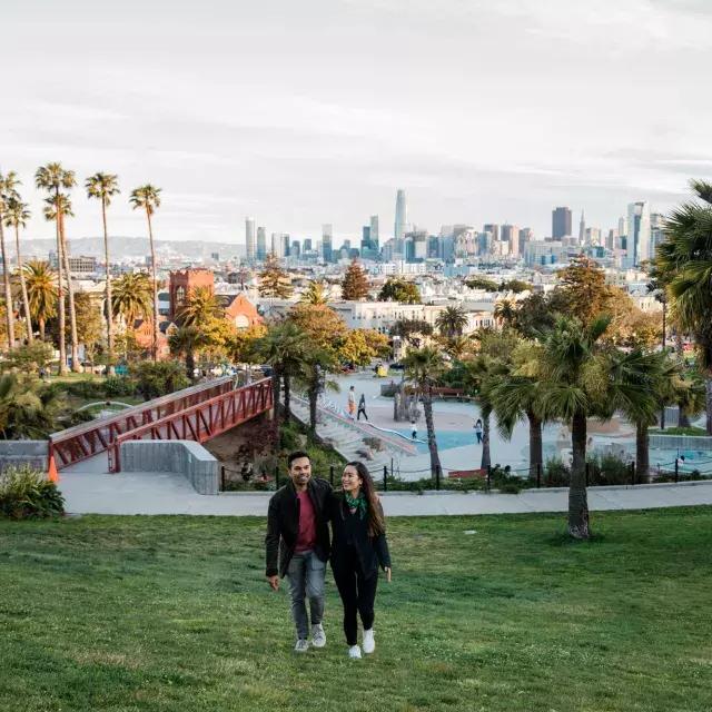 A couple walks toward the camera with 德洛丽丝公园 and the San Francisco Skyline behind them.